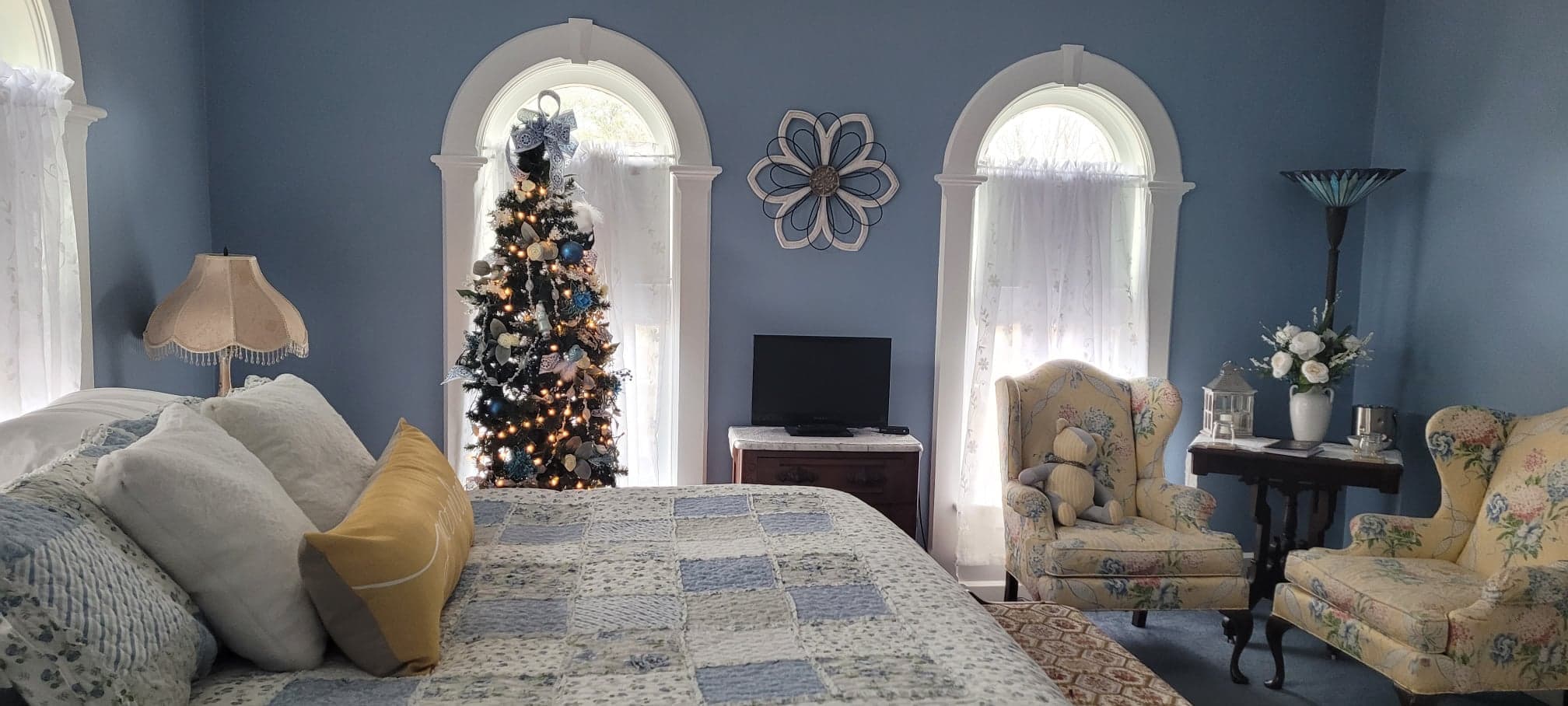 A panoramic view of a blue bedroom featuring arched windows flanking a lighted Christmas tree, a bed with a patchwork quilt on the left, and a sitting area with two floral wingback chairs on the right.