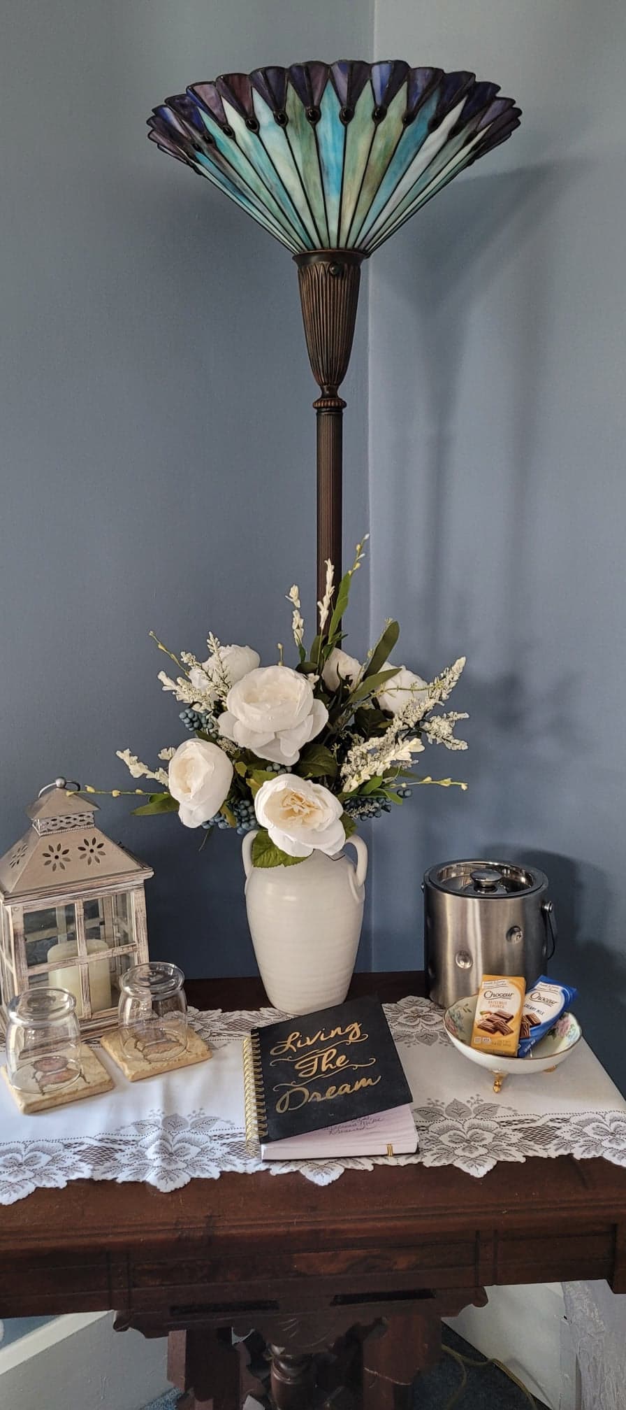 A welcoming corner display featuring a dark wood table holding a vase of white roses, a black 'Guest Book,' and a silver ice bucket, all illuminated by a tall Tiffany-style stained glass floor lamp.