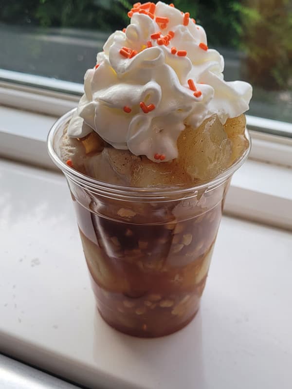 A clear plastic cup filled with layers of brown liquid, fruit, and topped with whipped cream and orange sprinkles.