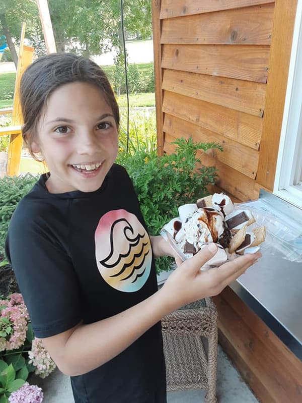 A smiling girl in a black shirt holds a plate stacked with ice cream and toppings outside a wooden building.