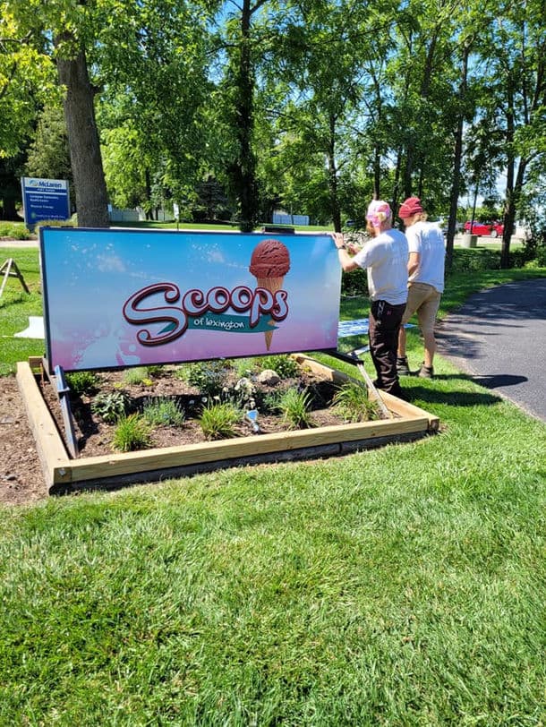 Two people are adjusting a colorful sign for "Scoops of Lexington" in a grassy area.