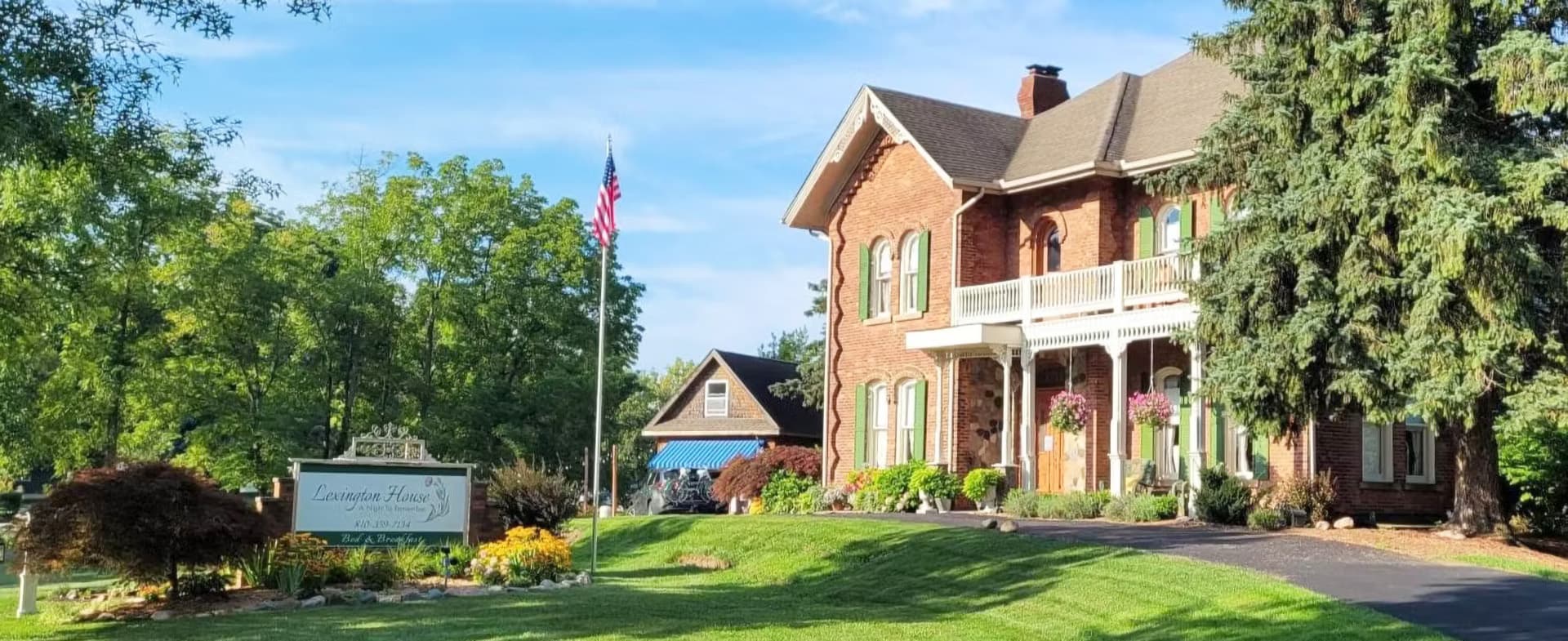 A charming brick house with a porch, surrounded by greenery and flowers, with an American flag and a sign for a business.