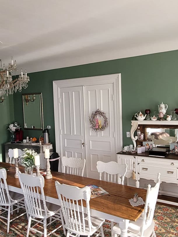 A cozy dining area featuring a long wooden table, white chairs, and a green wall adorned with decorative items.