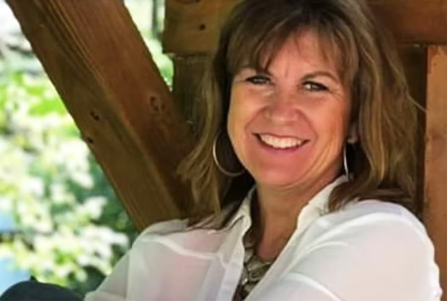 Smiling woman in a white shirt sitting on a wooden structure surrounded by greenery.